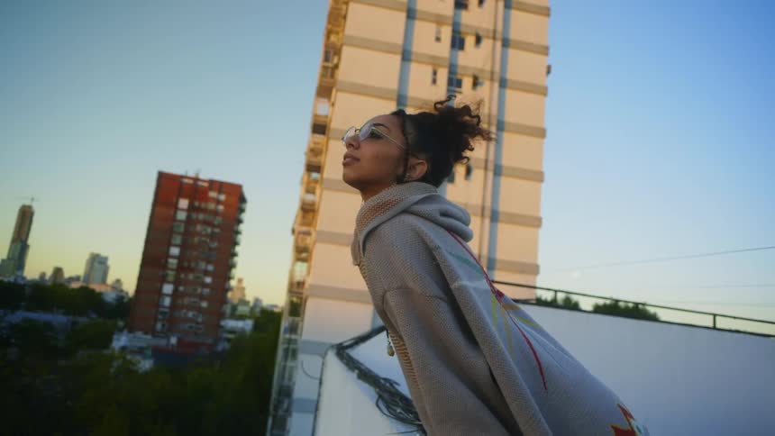 A young woman standing on a roof top leaning over a ledge with a tall building in the distance behind her