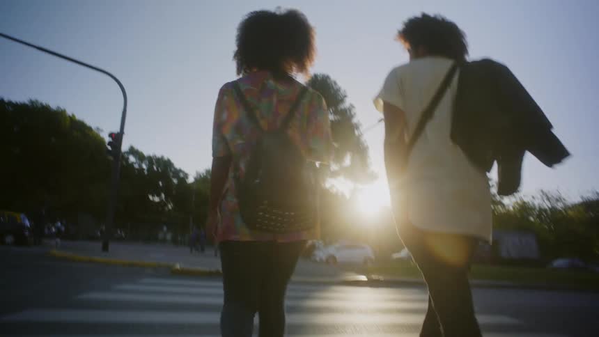 A man and woman walking across a street as sun shines through the trees