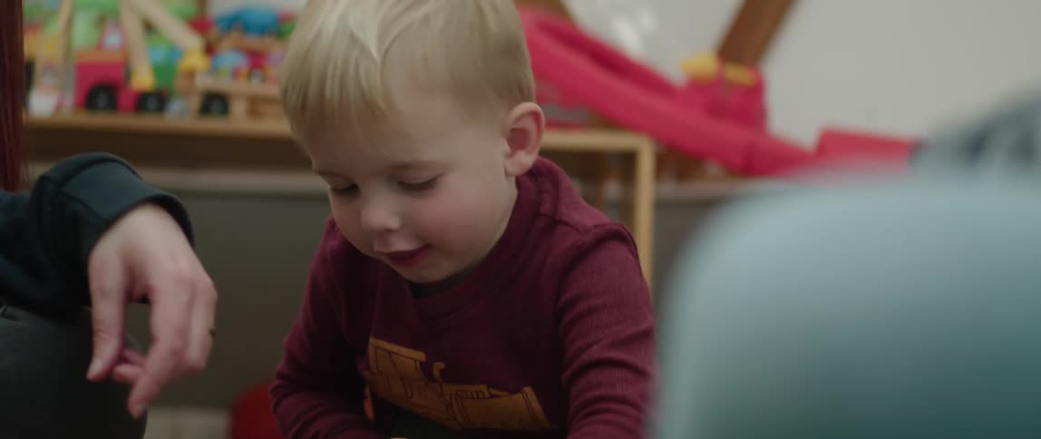 A toddler boy plays with a plastic truck while sitting on the floor near his mother and father
