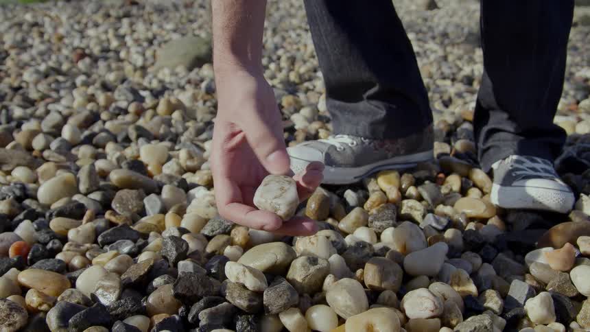 A man walking on pebbles and rocks leaning over and picking one up and looking it over in his hand