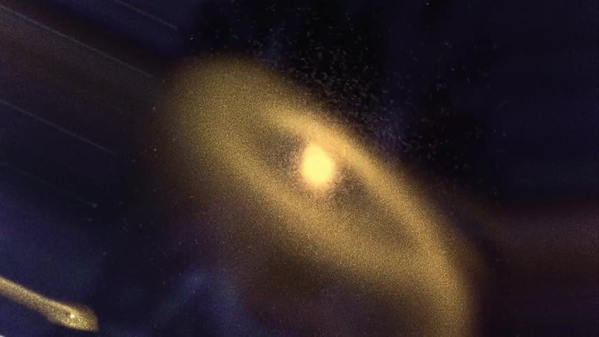 Glowing particles swirl around a rapidly rotating illuminated yellow sphere as soft colored lights move in the distance