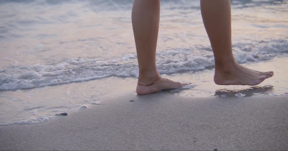 The lower legs of a barefoot person walking on a beach just at the waters edge during the daytime