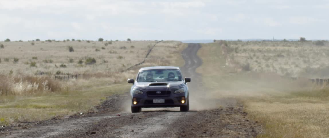 A car drives along a dirt road next to an open field
