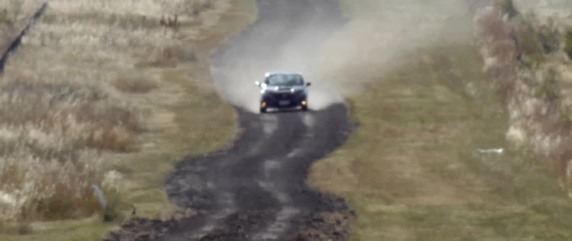 A dark sedan kicks up dust as it drives down a curvy dirt road through a field