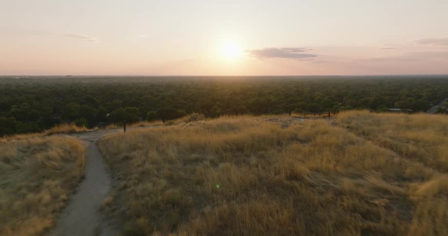 A bird flies over a neighborhood with houses and trees with high ground above and a dirt road in a rural area as the sun shines brightly on the horizon