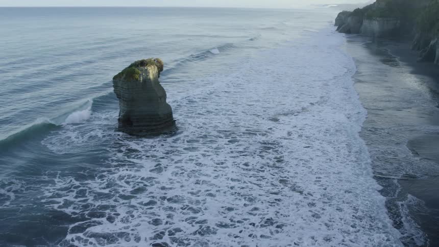 Waves roll into shore over a beach along a cliff as seagulls fly overhead and a rock formation stands in the water along the coast