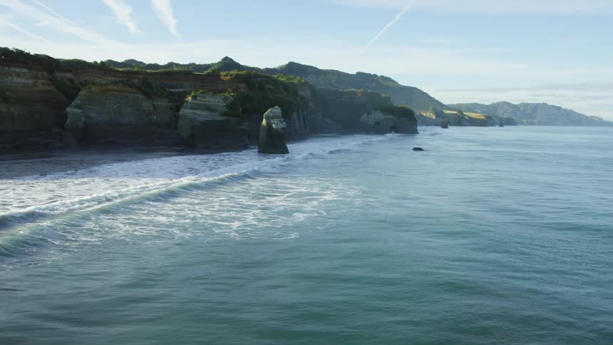 Waves roll in along the coast near a cliff with rock formations and mountains in the distance