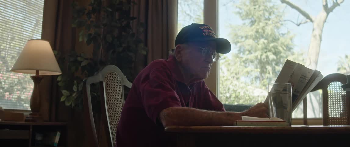 An elderly man sits at a dining table while reading a document next to a window