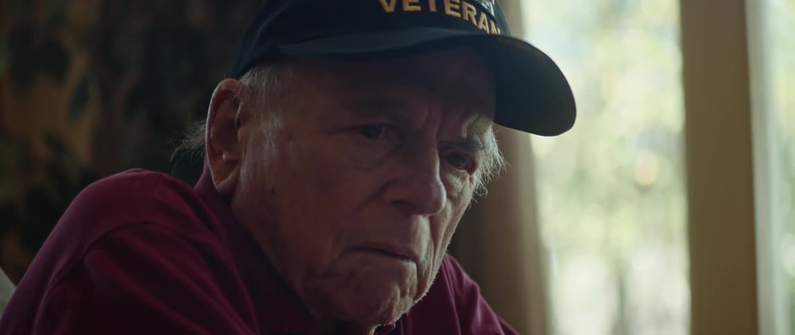 Elderly man with dark blue cap and crimson shirt takes off his glasses and shakes his head