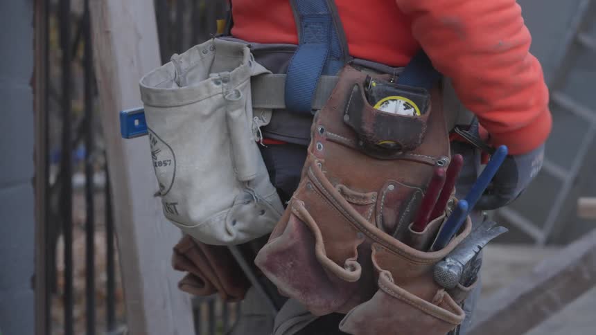A construction worker is standing outside as they reach back to the tools on their belt