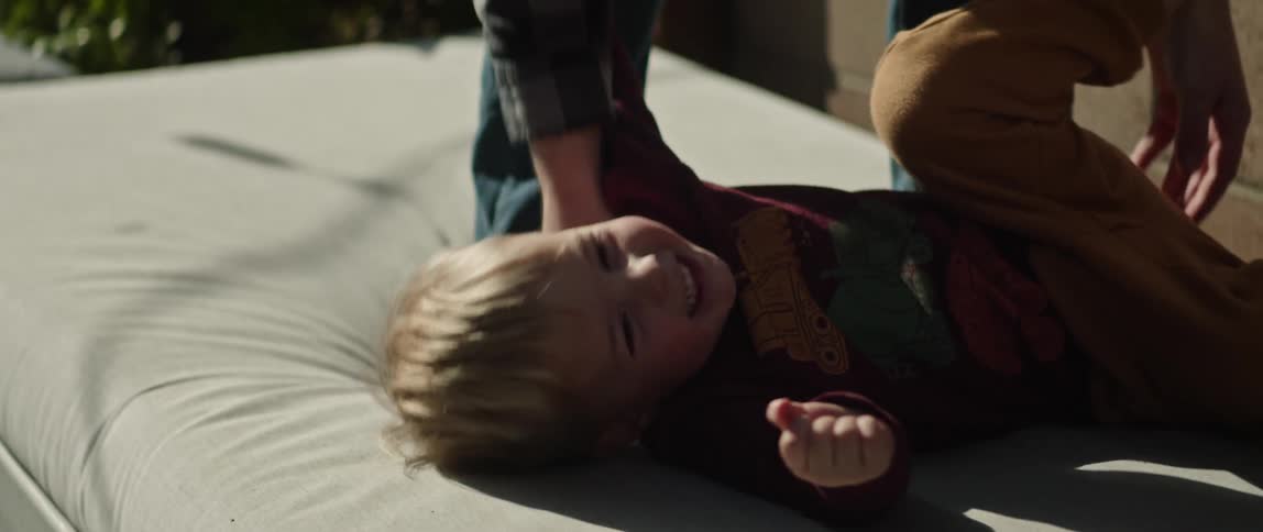 Father plays with son tickling the child as he lies on mattress outdoors near building