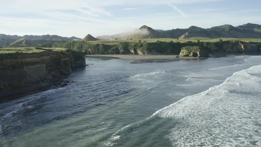 Aerial view from a drone of the rocky coastline of an ocean with waves hitting the shore and mountains in the distance