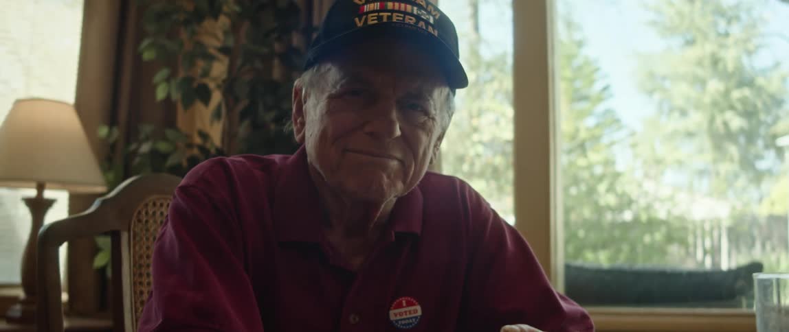 Elderly man with cap and crimson shirt smiles at camera in room