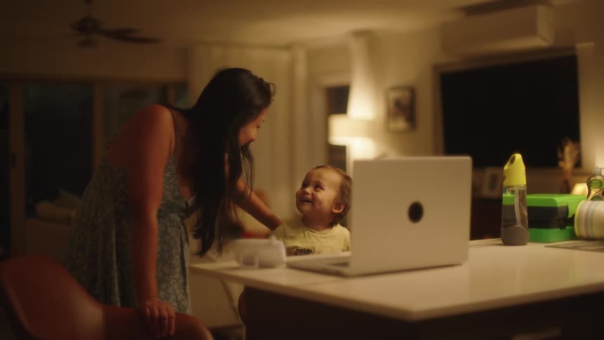 A mother and her young son are smiling at a laptop screen thats sitting on the kitchen countertop