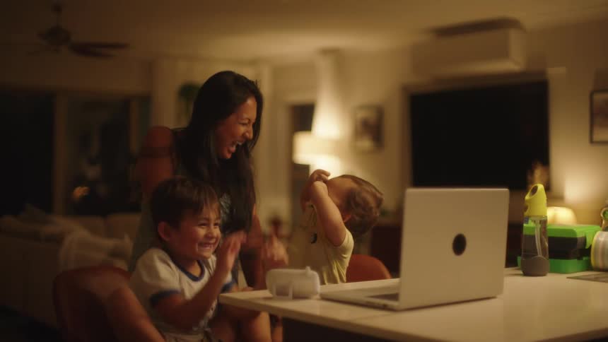 Happy family gathered on the living room smile and laugh at someone on a video call through a laptop