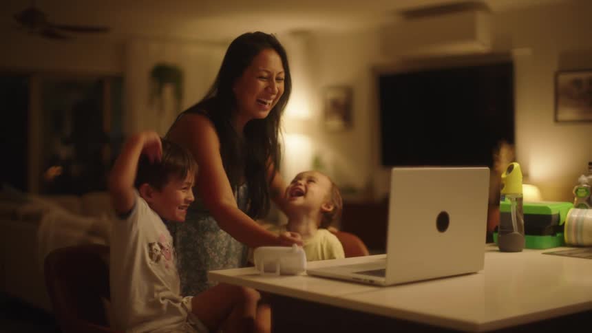 Two young boys sitting at a counter top laughing and smiling at a laptop while their mother walks over smiling and laughing and using the laptop