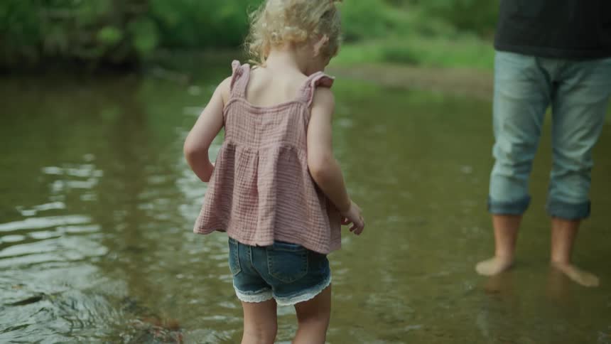 A little girl wearing shorts is walking in a shallow pond in the woods as her father is standing in the water watching