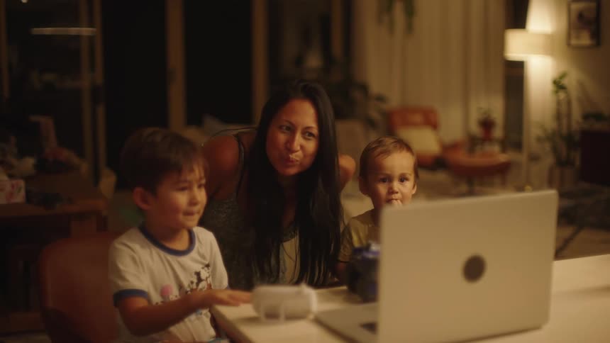 A mother leans over behind her two songs sitting watching a laptop screen while they laugh and smile
