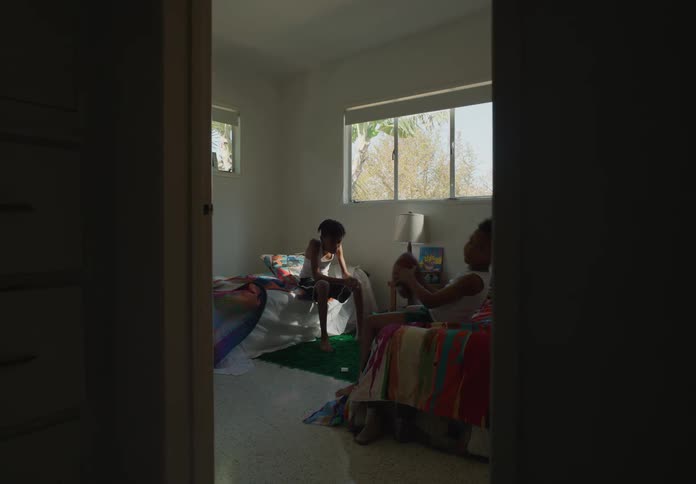 Two tween brothers in bedroom one sitting on edge of bed the other throwing football in the air