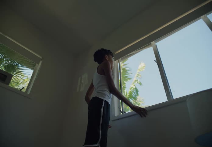A young boy standing in front of a window looking out as the wind blows the trees