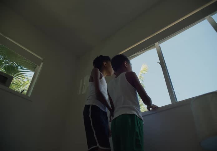 Two young boys stand together while looking out a bedroom window