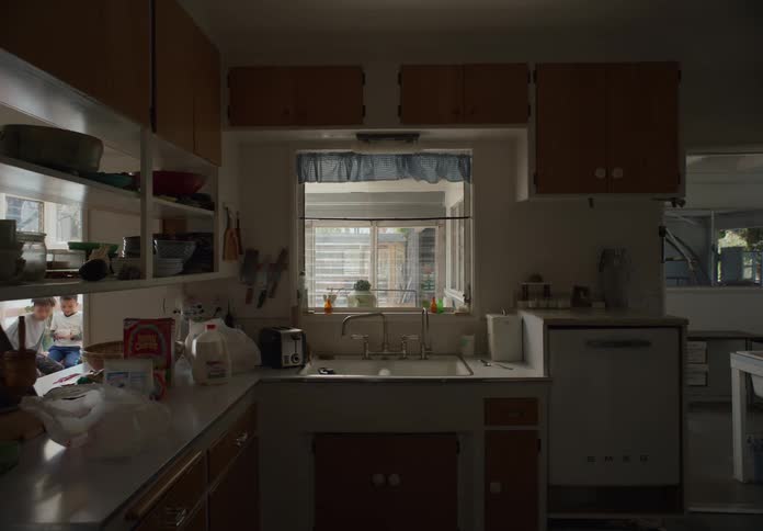 A boy standing in the kitchen washing his dishes while his brother waits in the adjoining sun room he joins him