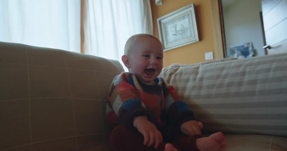 An infant is sitting upright on a couch indoors while laughing and smiling