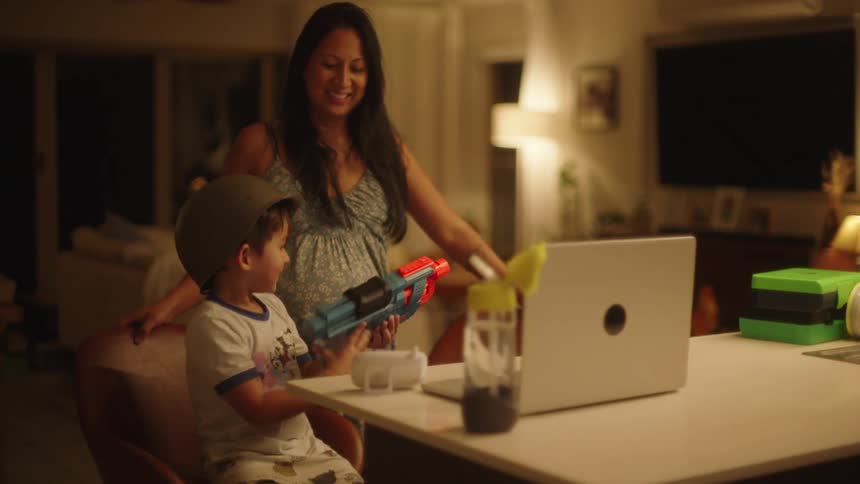 Pregnant mom and young son at dining table with laptop and toys smiling and laughing