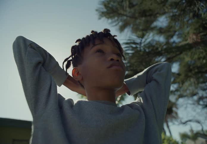 A young boy standing outside with his hand behind his neck looking ahead of him thinking