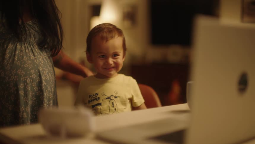 A little boy sits on a chair looking at a screen smiling and looking up at his mother enxt to him