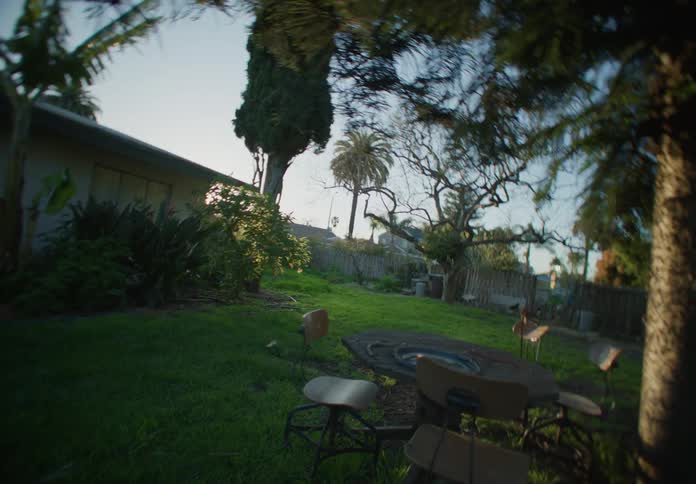 A back yard of a house with grass in the lawn and a table and chairs under a tree as the camera falls