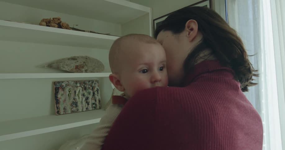 A woman holds a baby indoors in front of a bookshelf