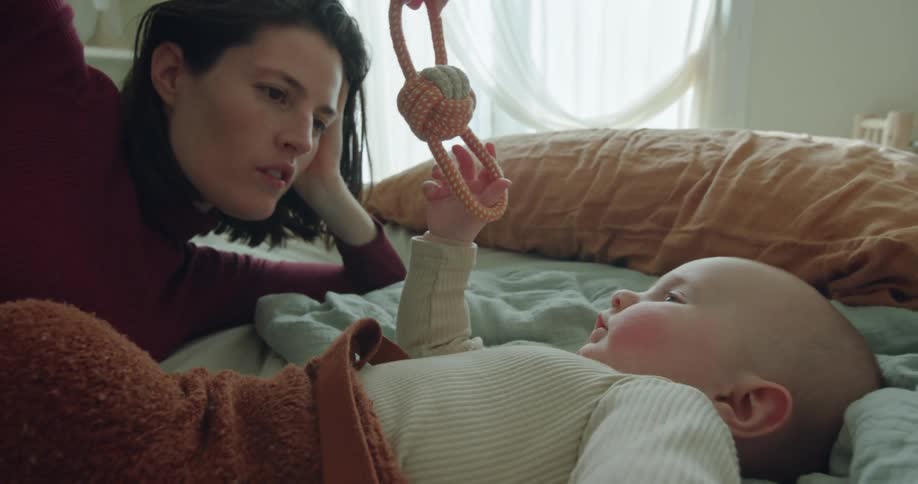 Mother laying in bed uses a toy to play with her baby