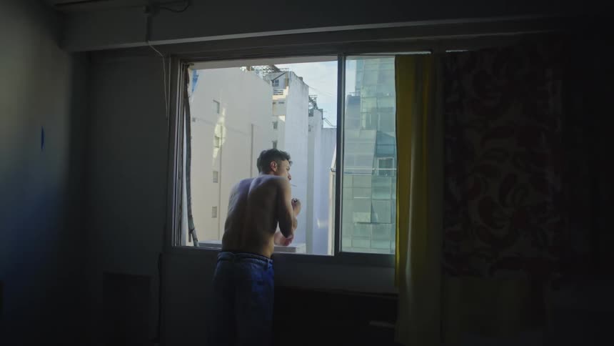 A man without a shirt on standing leaning out his window lighting a cigarette