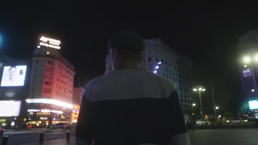 A man is walking on a busy downtown street at night as lights on the buildings glow in the darkness