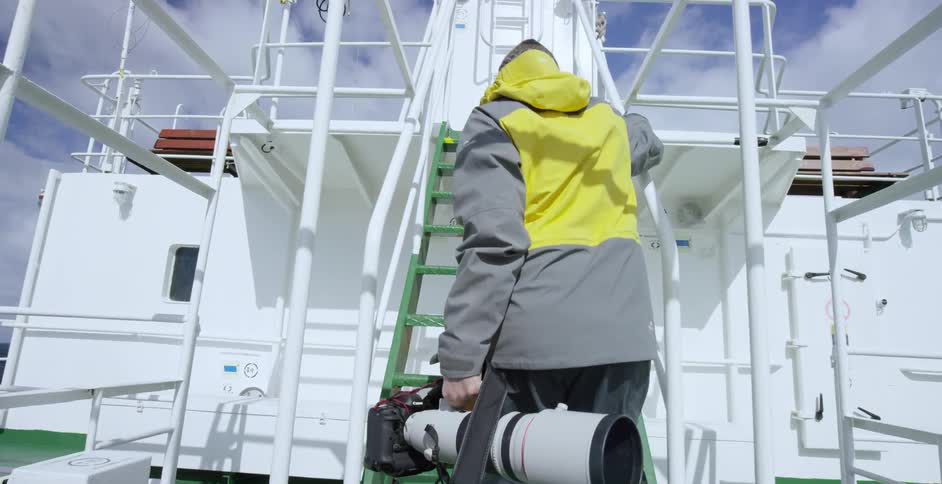 A man carries a large photo lens up the stairs of a boat