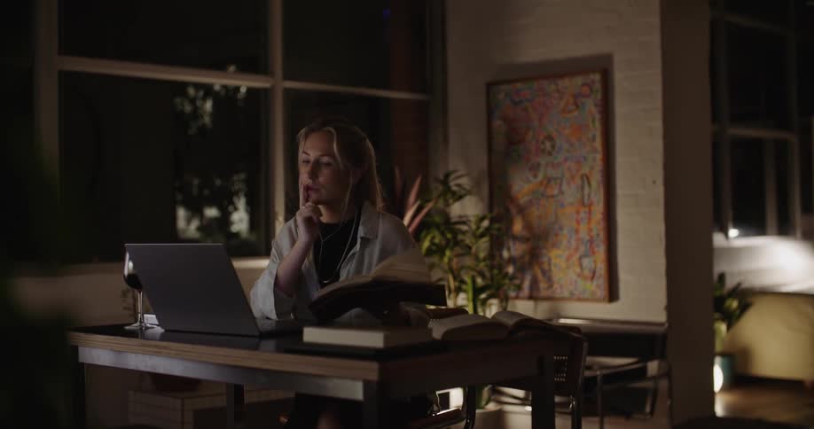 A woman reading a book at a desk ponders for a moment before beginning to type on a laptop indoors at night