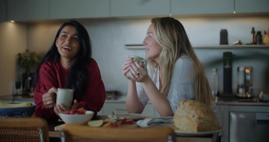 Two women in kitchen talking laughing and smiling while having coffee and brunch