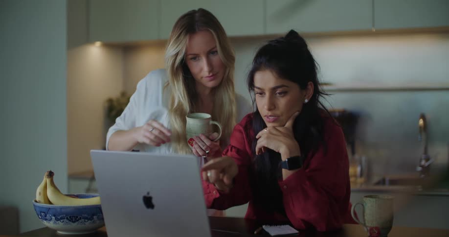 A woman is sitting at a table looking at a laptop computer as the woman standing next to her holding a cup of coffee points to the monitor