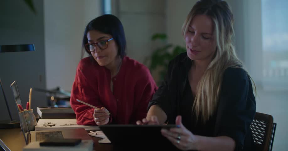Two young professional women have a conversation at an office desk while consulting a laptop and tablet