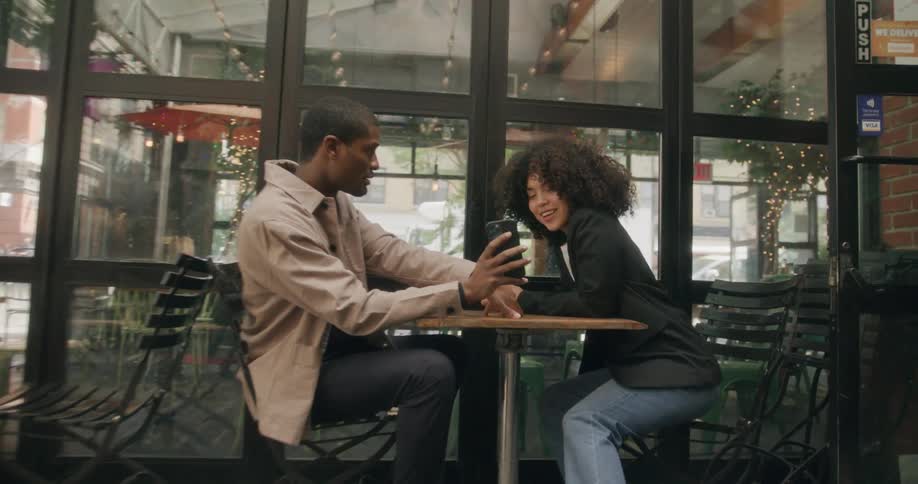 A man and woman sitting at a table outdoors looking at the mans phone in his hand