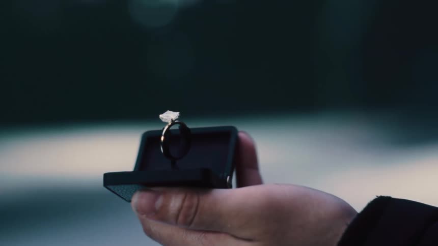 A hand holds a black ring box open to expose a fancy metal ring with a white stone