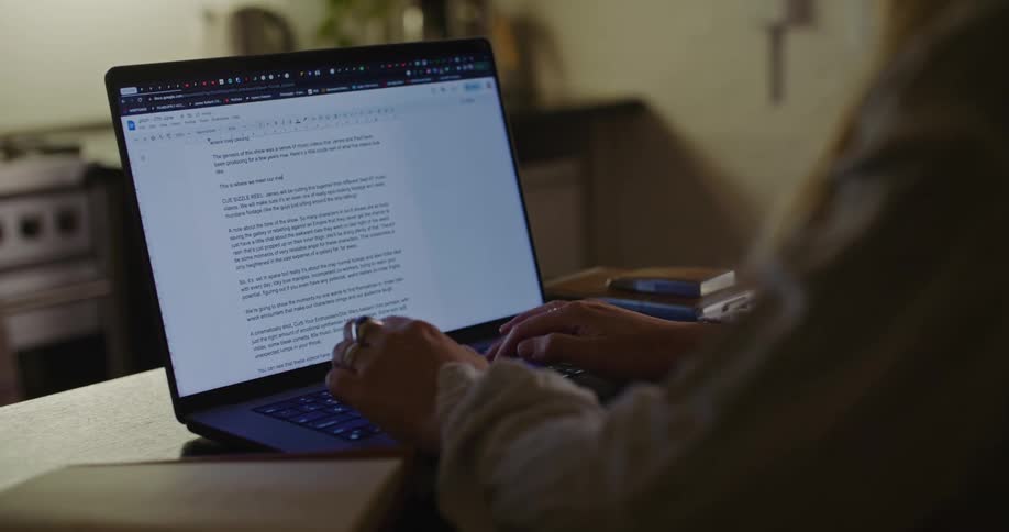 A woman at a kitchen table types a document using a laptop keyboard