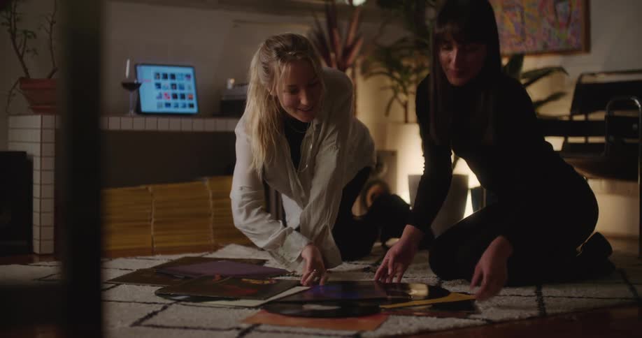 Two women are kneeling on a rug on the floor looking at vinyl record albums