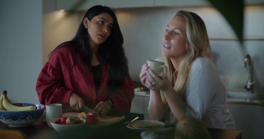 Two women are talking in a kitchen while one is chopping an apple