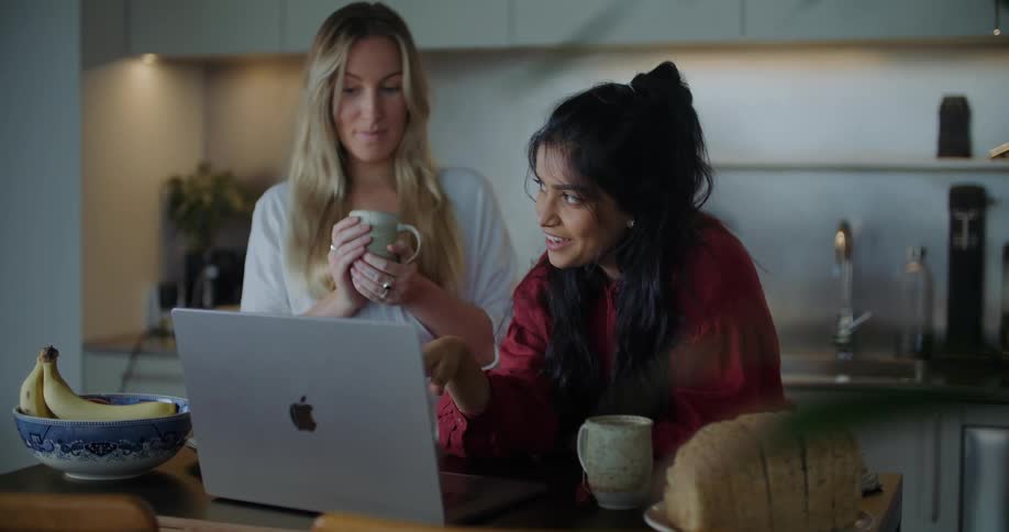 A woman is sitting at a table looking at a laptop computer as the woman standing next to her points to the monitor