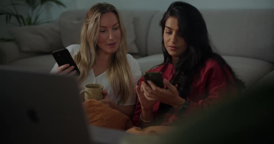 Two women are sitting on the floor in front of a table with a laptop computer as they look at their cell phones