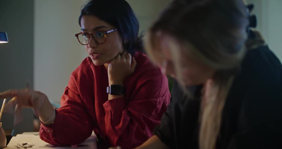Blonde woman with black shirt and woman with dark hair and red shirt sit at desk write in a notebook in room