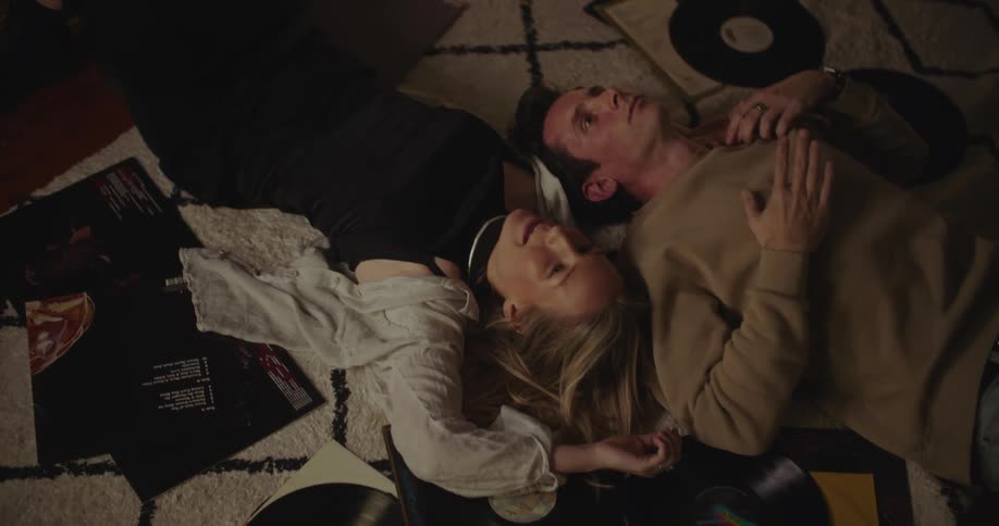 A man and woman have a conversation while laying on the floor and listening to music from the vinyl records nearby
