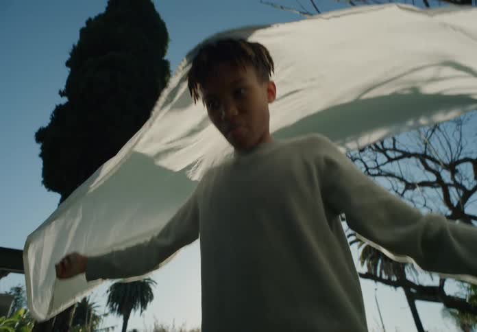 Boy swings on rope swing hung from tree in backyard as older brother holds bottom of sheet hanging on clothesline